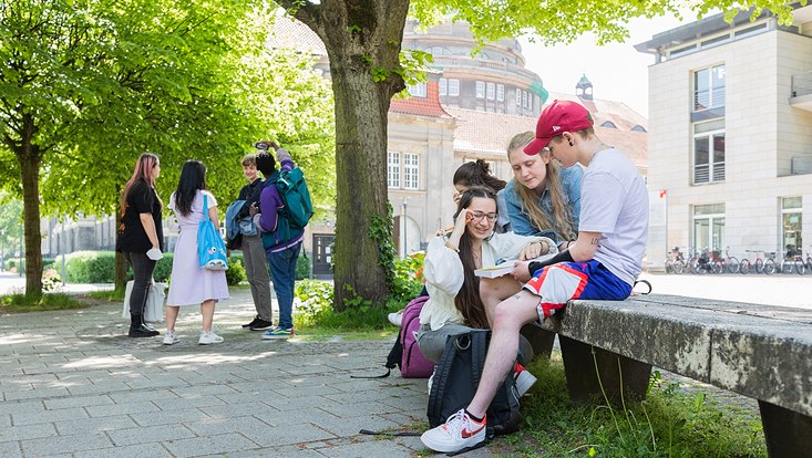 Rechts sitzen Studierende draußen bei schönem Wetter vor dem ESA und schauen in ein Buch. Links stehen Studierende und unterhalten sich.