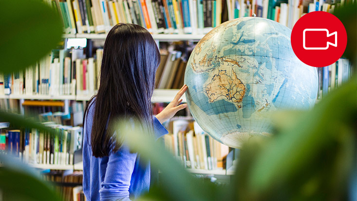 Blick auf eine Frau, die an einem Globus in einer Bibliothek steht. Oben rechts ist ein rotes Kamerasymbol.