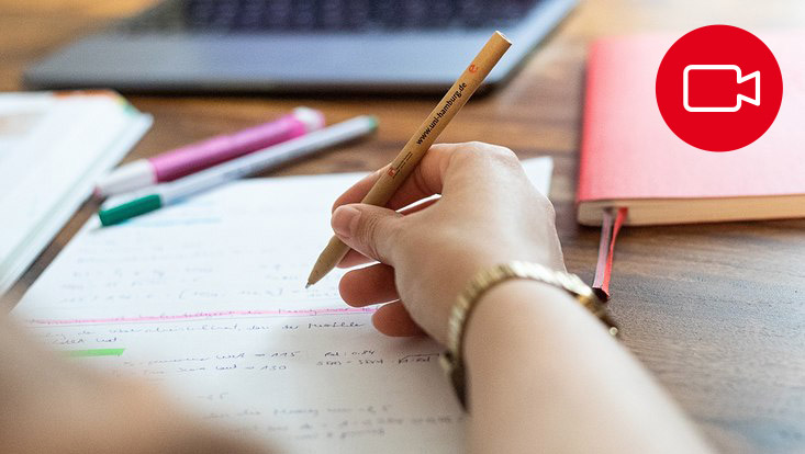 Eine Hand hält einen Stift und schreibt in ein Heft. Im Hintergrund liegen ein Buch, ein Laptop und Stifte.