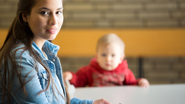 Eine Frau schaut in die Kamera und hinter ihr ist verschwommen ein Baby zu sehen.