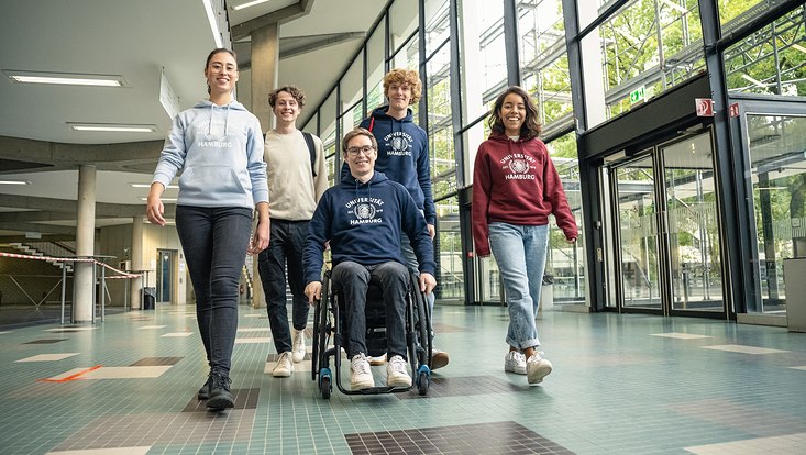 Fünf Studierende gehen im Audimax auf die Kamera zu. Einer von ihnen sitzt im Rollstuhl.