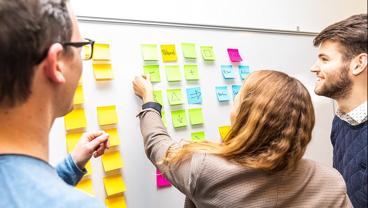 Drei Personen stehen vor einem Whiteboard mit Klebezetteln und arbeiten damit.