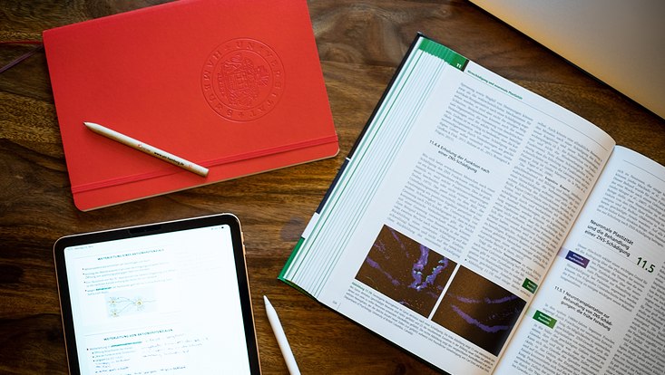 Blick auf ein offenes Buch, ein Tablet und ein Notizbuch.