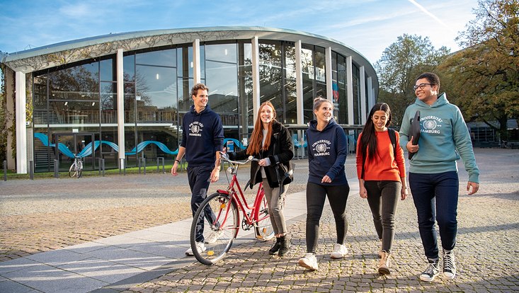 Blick auf fünf Studierende, die vor dem Audimax entlanggehen und sich unterhalten.