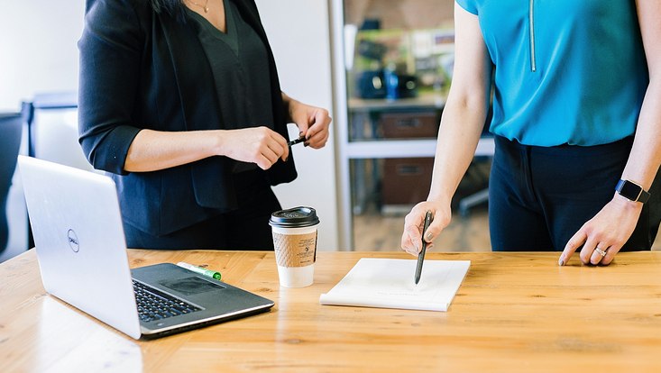 Im Vordergrund steht ein Schreibtisch, auf dem ein Laptop, ein Kaffeebecher und ein Notizblock liegen. An dem Tisch stehen zwei Personen, die sich unterhalten. Die rechte Person zeigt mit einem Stift etwas auf dem Notizblock.