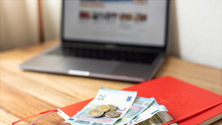 Ein Laptop, ein rotes Notizbuch mit Geldscheinen und Münzen liegen auf einem Tisch