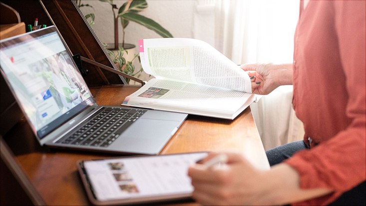 Blick auf eine Person, die online mit Laptop, Tablet und Buch studiert.