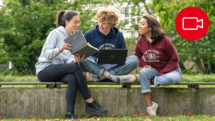 Drei Studierende sitzen draußen und unterhalten sich. Eine Person hält einen Laptop und eine andere hält ein Notizbuch.