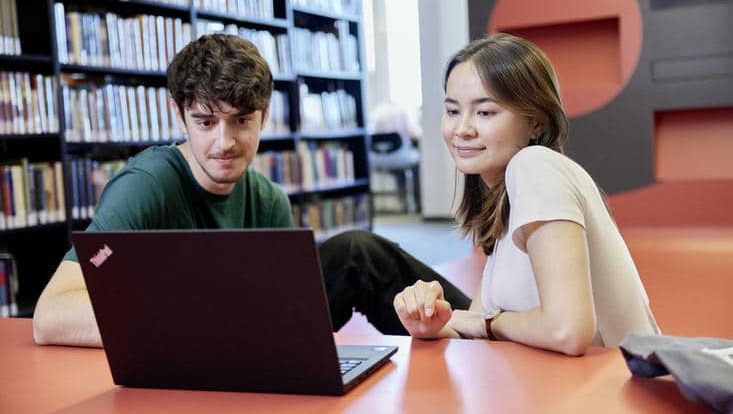 Zwei Studierende sitzen in der Bibliothek gemeinsam vor einem Laptop